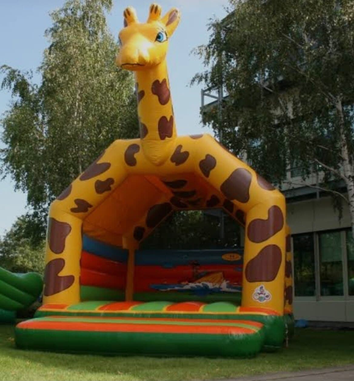 Gelbe Giraffen-Hüpfburg mit braunen Flecken, der lange Hals spannt sich über den Eingang, auf einer Wiese